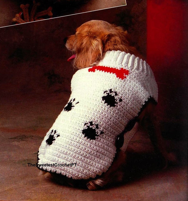 Puede incluir: Un su&eacute;ter de ganchillo blanco para perro con cuello acanalado, un motivo rojo en forma de hueso cerca del escote y patrones de huellas negras en la espalda. La prenda tiene un borde negro en la parte inferior e incluye el texto TheSweetestCrochetPT.