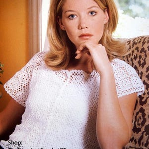 May include: A white short-sleeved crochet top featuring an intricate openwork pattern with a scalloped neckline and hem. The garment has a lightweight, airy construction with a delicate, repeating geometric stitch design throughout the fabric.