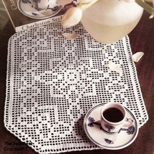 May include: White crocheted doily with a scalloped edge, featuring a delicate floral pattern. A white cup and saucer with a blue floral design sits on top of the doily.