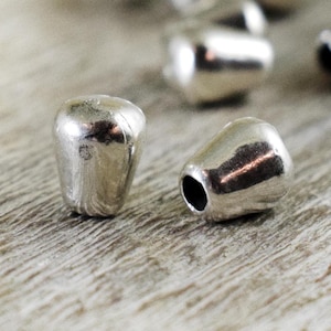 May include: Two silver metal bead caps, each with a rounded, cone-shaped top. The caps are lying on a light brown wooden surface.
