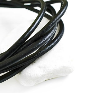 Può includere: Primo piano di un cordoncino in pelle nera, arrotolato e appoggiato su una superficie bianca.