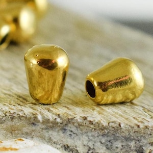 Puede incluir: Dos cuentas de metal de color dorado, una con la parte superior redondeada y la otra con la parte superior puntiaguda, se muestran sobre una superficie de madera clara.