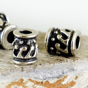 Könnte beinhalten: Silberne Metallperlen mit einem komplizierten Design. Die Perlen sind zylindrisch und haben eine glatte, polierte Oberfläche.