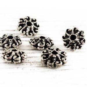 May include: Six silver-toned metal bead caps with a decorative floral design. The caps have a hole in the center for threading.