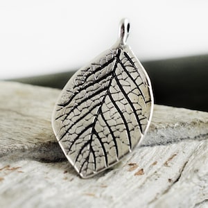 Puede incluir: Un dije de hoja de plata con un diseño texturizado detallado. El dije tiene un bucle en la parte superior para sujetarlo a un collar o pulsera.