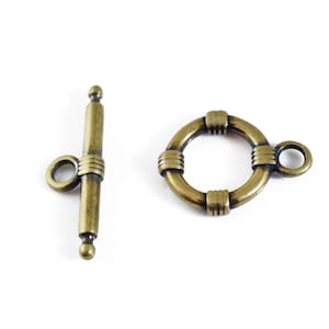 May include: A brass-toned metal toggle clasp set consisting of a T-bar component with a central loop and a circular ring component featuring three decorative ribbed sections and an attached eyelet for jewelry assembly and fastening.