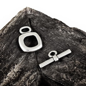 May include: Two silver-coloured jewellery clasps. One is a square shape with a loop at the top, and the other is a bar with a ring on one end. The clasps are on a dark, textured surface.