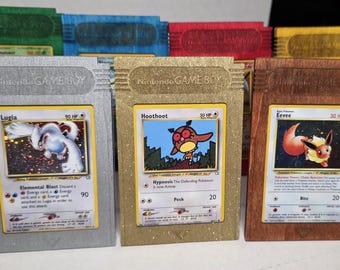 Könnte beinhalten: Eine Sammlung von maßgefertigten Nintendo Game Boy-Kassettenhaltern, die jeweils eine Pokémon-Sammelkarte zeigen. Die Halter sind in verschiedenen Farben erhältlich, darunter Silber, Gold und Holzfarben, mit dem Nintendo Game Boy-Logo.