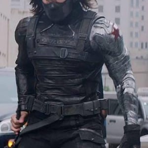 May include: A person in a black tactical outfit, featuring a mask, gloves, and a metal arm. The outfit includes a chest plate, belt, and black trousers. A red star is visible on the metal arm.