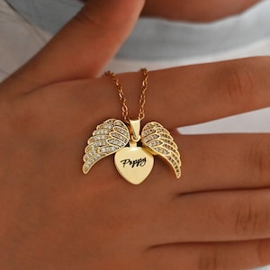 Pode incluir: Colar dourado com um pingente em forma de coração e detalhes de asas. O pingente tem o nome "Poppy" gravado. As asas são incrustadas com pequenas pedras transparentes.