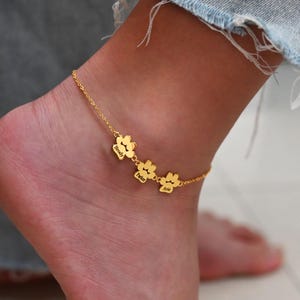 Puede incluir: Una tobillera dorada con tres colgantes en forma de huella de pata, cada uno grabado con un nombre. La tobillera se lleva en un tobillo, con unos vaqueros en el fondo. Los nombres en los colgantes son Brody, Baker y Arie.