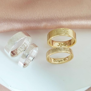 Op de afbeelding: Twee gepersonaliseerde vingerafdrukringen. Een ring is goud met de namen "Kelly" en "Georgie" gegraveerd. De andere ring is zilver met de datum "1.13.23" gegraveerd.