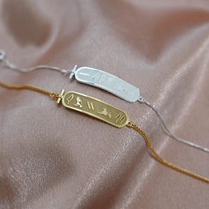 May include: Two delicate bracelets with rectangular pendants featuring ancient Egyptian hieroglyphs. One bracelet is gold-toned, the other silver-toned, both resting on a pink satin surface. The bracelets have a chain-link design.
