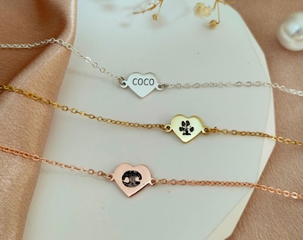 May include: Three silver heart-shaped necklaces with different engravings. The top necklace has the word "Coco" engraved on it. The middle necklace has a paw print engraved on it. The bottom necklace has a heart engraved on it.