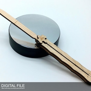 Puede incluir: Un cuchillo mariposa de madera clara, cortado con láser, descansa sobre una superficie reflectante. El cuchillo está en posición abierta, mostrando su intrincado diseño. Las palabras "DIGITAL FILE" están en la parte inferior.