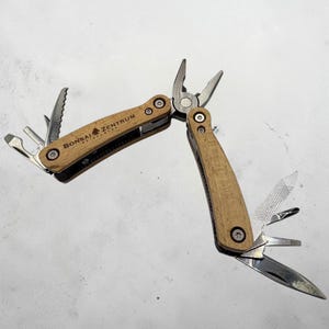 Könnte beinhalten: Ein Multitool mit hellem Holzmaserungsgriff und verschiedenen Werkzeugen, darunter eine Zange, eine Säge und ein Messer. Der Text "BONSAI ZENTRUM" ist auf dem Griff aufgedruckt. Das Werkzeug ist geöffnet und zeigt seine verschiedenen Komponenten.
