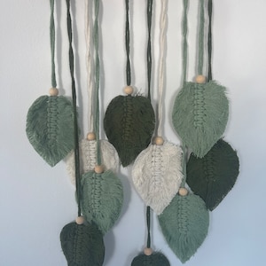Könnte beinhalten: Ein Wandbehang aus Makramee mit mehreren grünen und cremefarbenen Blättern. Die Blätter sind aus Garn gefertigt und haben unten Holzperlen.