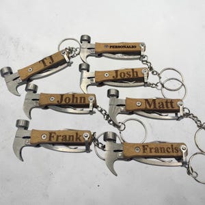Könnte beinhalten: Eine Sammlung von schlüsselanhängern in Hammerform. Jeder Schlüsselanhänger hat einen Holzgriff, einen silberfarbenen Hammerkopf und ein Klappmesser. Namen wie "John", "Josh" und "Frank" sind in das Holz graviert.