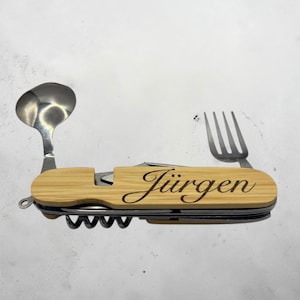 Könnte beinhalten: Ein Multi-Tool mit Bambusgriff, mit Löffel, Gabel, Messer und Korkenzieher. Der Griff ist mit dem Namen "Jürgen" graviert. Das Werkzeug ist silber- und bambusfarben.