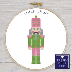 May include: A cross-stitch pattern of a nutcracker soldier in a wooden embroidery hoop. The nutcracker is pixelated with green, pink, gray, and white colors. The words "stitch chart" are written in blue at the top. A logo is in the bottom right corner.
