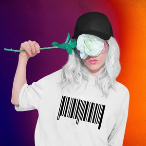 Könnte beinhalten: Ein weißes T-Shirt mit einem schwarzen Barcode auf der Vorderseite. Die Person trägt eine schwarze Baseballkappe und eine weiße Rose bedeckt ihr Gesicht.