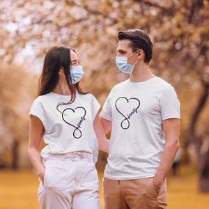 May include: Two people wearing white t-shirts with a black infinity symbol and the word "family" inside a heart. They are both wearing blue face masks and standing in a park with fall foliage in the background.