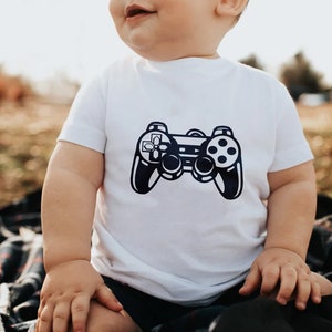 Pode incluir: Uma camiseta branca para bebê com um gráfico preto de um controlador de videogame.