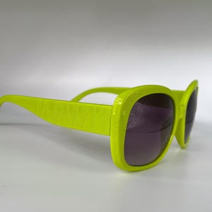 Może przedstawiać: Jasne neonowo-zielone okulary przeciwsłoneczne z teksturowanym wzorem na zausznikach i ciemnofioletowymi soczewkami. Okulary przeciwsłoneczne mają kwadratową oprawkę i odważny, nowoczesny design, idealny do dodania koloru do każdego stroju.