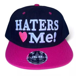 Puede incluir: Gorra snapback negra y fucsia con el texto "HATERS ❤️ Me!" en blanco. La gorra tiene una visera plana y un corazón rosa. La gorra forma parte de la colección R-M Sport Cap & Hat.