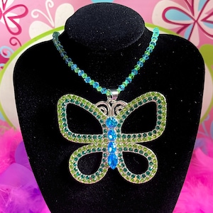 May include: A necklace featuring a large butterfly pendant encrusted with green and blue rhinestones. The butterfly body is made of bright blue teardrop-shaped stones. The chain consists of alternating small green and blue faceted crystal beads.