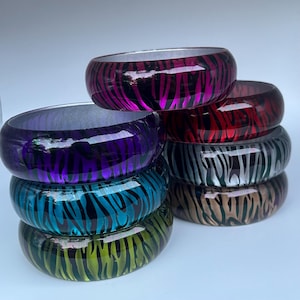 May include: Stack of colorful, wide bangles with a zebra stripe pattern. The bangles are in shades of purple, teal, green, red, silver, brown, and pink. The bracelets are made of a glossy material.