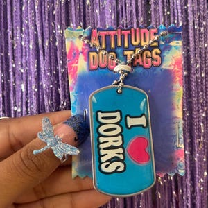 May include: A blue metal dog tag necklace on a tie-dye display card labeled ATTITUDE DOG TAGS. The tag features the text I HEART DORKS with a pink heart graphic. A silver ball chain connects the tag to the card, which has a scalloped top edge.