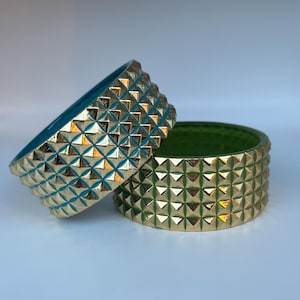May include: Two decorative bowls with a geometric pattern. One bowl is teal with gold accents and the other is green with gold accents. The bowls have a textured, studded exterior and a smooth interior.