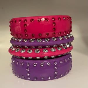 May include: Stack of three colorful bangles. The top bangle is hot pink with small, red rhinestones. The middle bangle is also pink, with clear rhinestones. The bottom bangle is purple with purple rhinestones.