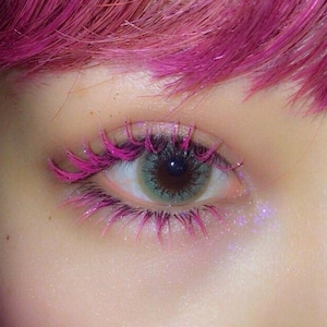 Pode incluir: Close-up de um olho com cílios rosa e lentes de contato verdes.