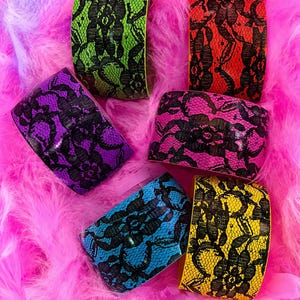 Può includere: Sei bracciali a fascia larga con un motivo floreale in pizzo nero sovrapposto a vivaci sfondi neon color verde lime, rosso, viola, rosa acceso, blu brillante e giallo. Ogni bracciale ha una superficie liscia e curva dal design ad alto contrasto.
