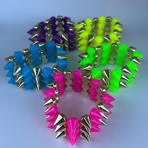 Può includere: Quattro braccialetti a punte colorati. I braccialetti sono nelle tonalità di viola, giallo neon, verde neon, blu e rosa. Ogni braccialetto ha punte color oro e una base abbinata. I braccialetti sono disposti su una superficie bianca.