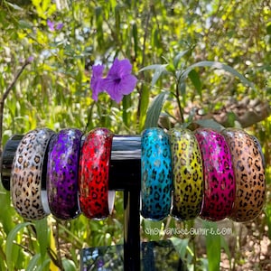 May include: Seven colorful bangle bracelets with a leopard print design. The bracelets are arranged in a row on a black stand. The bracelets are made of a hard plastic material and have a glossy finish.