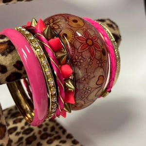 Op de afbeelding: Een verzameling gestapelde armbanden in verschillende stijlen en kleuren. De armbanden omvatten een felroze armband, een goudkleurige armband met heldere strass-steentjes en een bruine armband met bloemenpatroon. Sommige armbanden hebben gouden spikes en roze accenten.