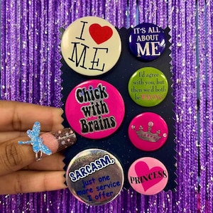May include: A collection of seven circular pin-back buttons displayed on a dark blue card. The buttons feature various colors including white, dark blue, green, and pink, with text such as I Love Me, It's All About Me, Chick with Brains, and Sarcasm.