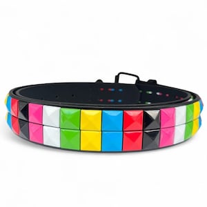 Pode incluir: Um cinto preto com tachas quadradas em forma de pirâmide coloridas. As tachas estão dispostas num padrão repetitivo de azul, vermelho, rosa, branco, verde, amarelo e preto. O cinto tem uma fivela preta e pequenos pontos coloridos.