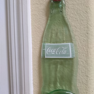 May include: A green glass Coca-Cola bottle-shaped wall hanging with a red bottle cap. The Coca-Cola logo is visible on the label. The bottle is mounted on a light yellow wall next to a white trim.