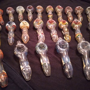 Puede incluir: Una colección de 20 pipas para fumar de vidrio con varios diseños y patrones coloridos. Las pipas están hechas de vidrio transparente y tienen una cazoleta redonda y una boquilla larga y delgada.