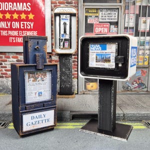 Może przedstawiać: Miniaturowy niebieski automat z gazetami Daily Gazette oraz biały automat USA News stojące na chodniku. Obie jednostki posiadają przezroczyste okienka z miniaturowymi gazetami oraz szczegółową konstrukcję przypominającą metal.
