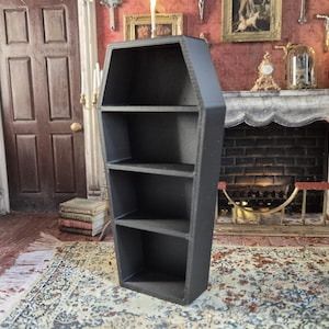 Puede incluir: Una estantería negra en forma de ataúd con tres estantes. La estantería es de madera y se mantiene en posición vertical. El fondo incluye una chimenea, una figura de hombre y una puerta.