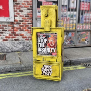 Op de afbeelding: Een miniatuur gele krantenautomaat met een verweerde afwerking. Het displayvenster toont een krant met een clownafbeelding en de tekst STOP THE INSANITY. Het onderste paneel toont de tekst NEW YAWK POST in dikgedrukte zwarte letters.