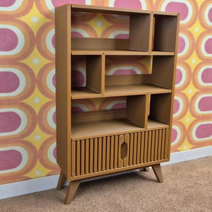 May include: A mid-century modern style wooden bookcase with multiple shelves and a lower cabinet. The bookcase is a warm brown color, with a vertical slatted design on the cabinet door. It stands on angled legs.