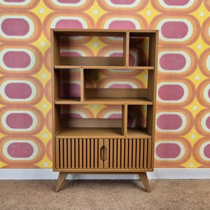 May include: A light brown wooden bookshelf with multiple compartments and a lower cabinet with vertical slats. The bookshelf has a mid-century modern design, with angled legs and a handle on the cabinet door. The background features a retro-style wallpaper.
