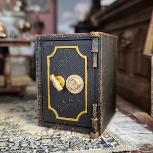 May include: A miniature black safe with gold detailing. The safe features a textured surface and a round lock. The scene includes a bust, books, and other decorative items, suggesting a vintage or antique theme.