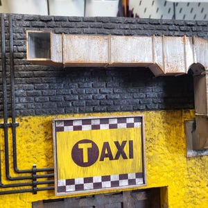 Może przedstawiać: Żółto-czarny znak w szachownicę z napisem "TAXI" białymi literami i czarnym kółkiem z białym "T" w środku. Znak jest zamontowany na ceglanej ścianie z metalową rurą wentylacyjną nad nim.